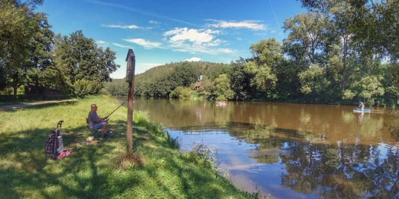 Man fishing by a calm riverside, surrounded by trees and greenery, under a clear sky at Prima