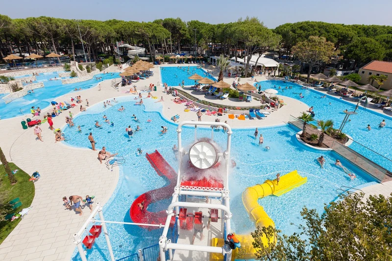 People swimming and playing in large, interconnected pools with slides and sunbathing areas surrounded by lush trees at Ca'Pasquali