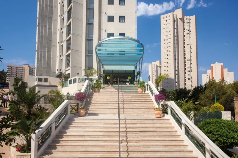 A wide staircase with plants ascends to a building entrance with a glass canopy; tall buildings surround in an urban area at Benidorm Horizon