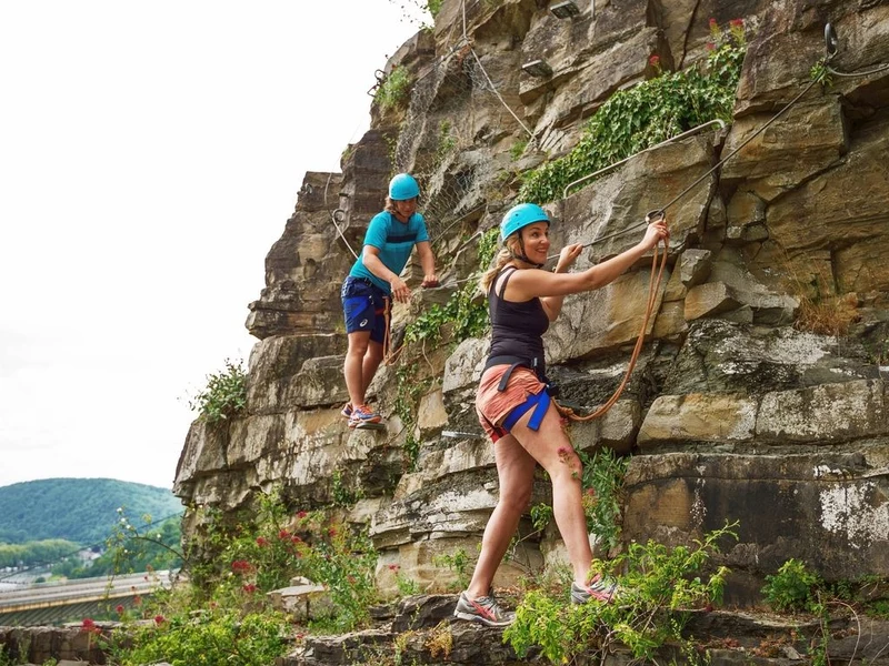 Two people climbing a rocky wall wearing helmets and harnesses, amid green vegetation at Landal Village les Gottales