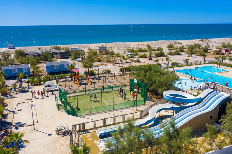 Swimming pools, slides, and sports court amidst trees, people, and mobile homes near the beach at Marvilla Parks Le Palavas